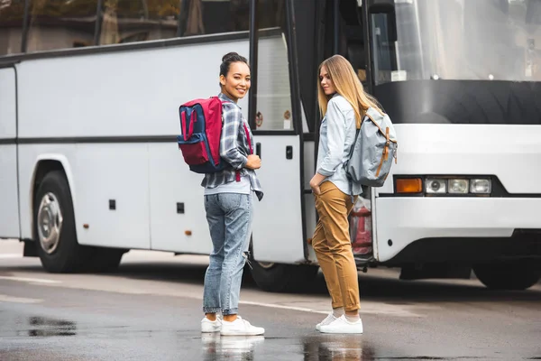 Молодые мультиэтнические женщины с рюкзаками позируют возле туристического автобуса на городской улице — стоковое фото