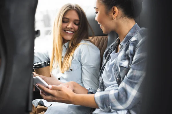 Selective focus of asian woman showing smartphone to her female friend with paper cup of coffee during trip on bus — Stock Photo