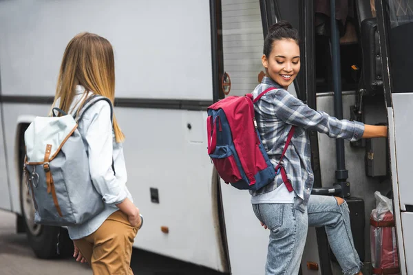 Young multiethnic female travelers with backpacks walking into travel bus at street — Stock Photo