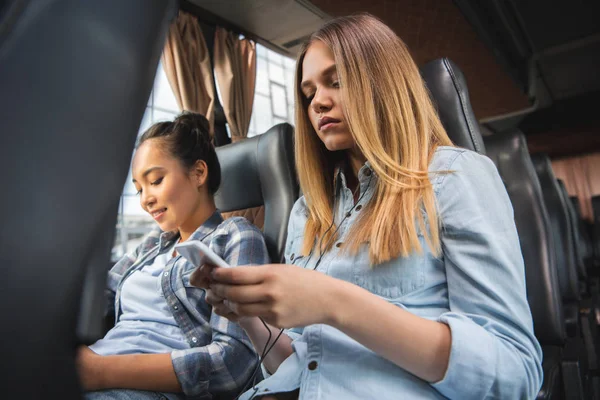 Приваблива жінка сидить зі смартфоном, а її азіатська подруга сидить поруч під час подорожі на автобусі — стокове фото