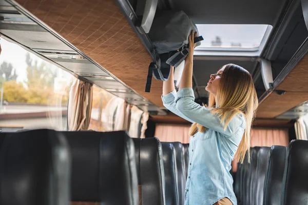 Side view of young woman putting backpack on shelf in travel bus — Stock Photo
