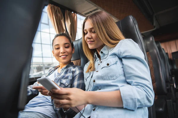 Feliz joven mujer mostrando smartphone a su asiático hembra amigo durante viaje en autobús - foto de stock