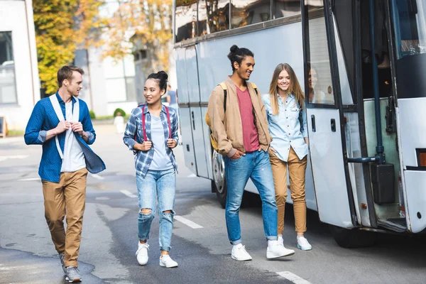 Smiling multicultural friends with backpacks walking near travel bus at street — Stock Photo