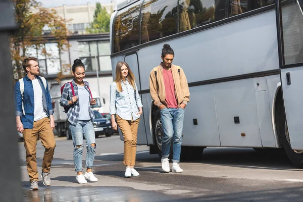 Selective focus of multiethnic friends with backpacks walking near travel bus at urban street — Stock Photo