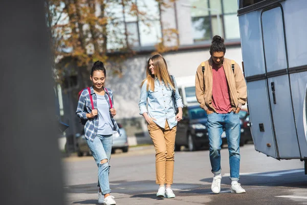 Enfoque selectivo de jóvenes amigos multiétnicos con mochilas caminando cerca de autobús en la calle de la ciudad - foto de stock
