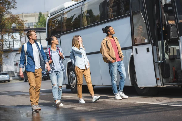 Cheerful multiethnic tourists with backpacks walking near travel bus at street — Stock Photo