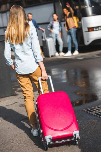 Vista trasera de la mujer que lleva la bolsa de ruedas mientras sus amigos esperan cerca de autobús de viaje en la calle - foto de stock
