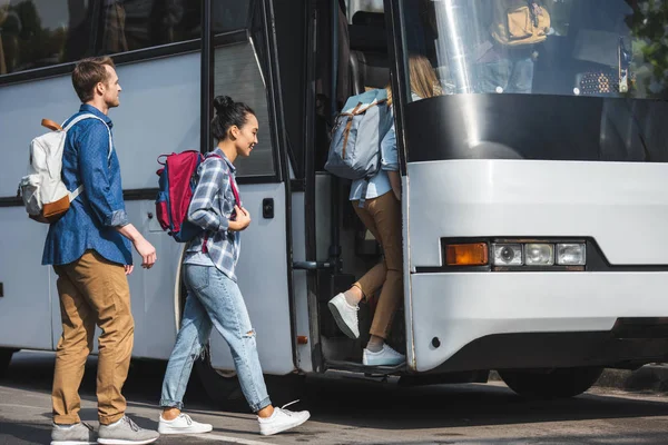 Веселі багатоетнічні друзі з рюкзаками, що йдуть в туристичний автобус на міській вулиці — стокове фото