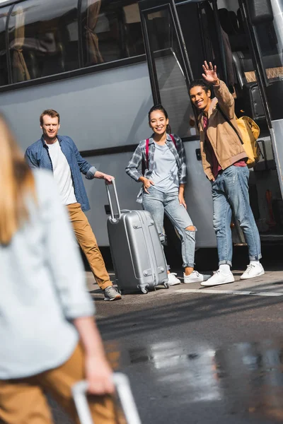 Sonriente joven de raza mixta saludando a mano mientras está de pie con amigos multiculturales cerca de autobús de viaje en la calle - foto de stock