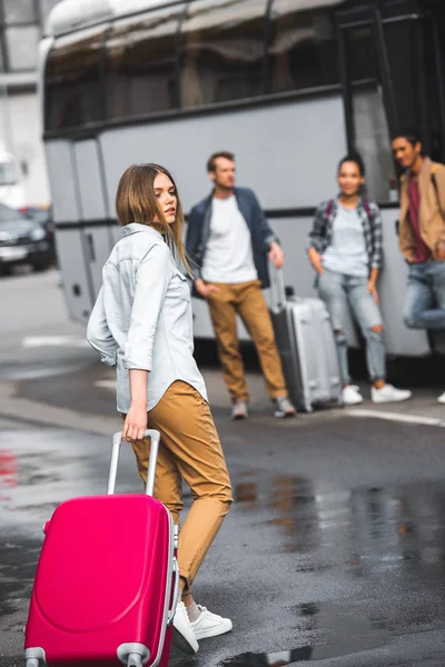 Красивая туристка с колёсной сумкой в то время как ее друзья ждут возле автобуса на городской улице — стоковое фото
