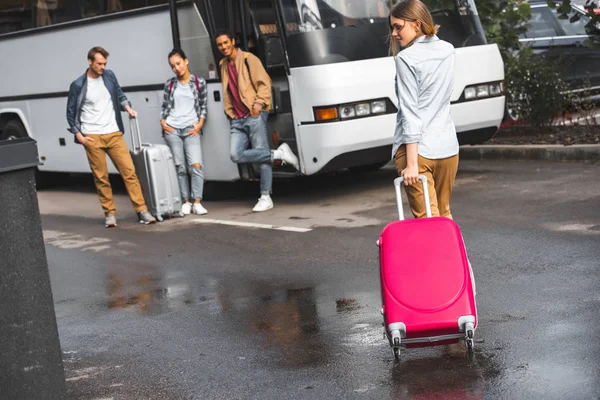 Rückansicht der attraktiven Frau mit Rolltasche, während ihre Freunde in der Nähe des Reisebusses auf der Straße warten — Stockfoto