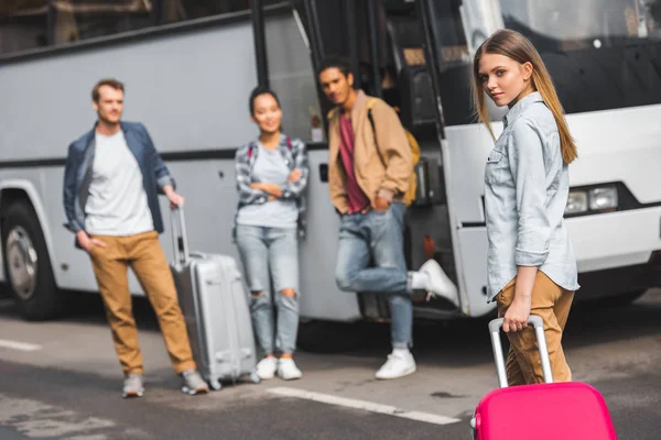 Selektiver Fokus der jungen Frau mit Rolltasche, während ihre Freunde in der Nähe des Reisebusses auf der Straße warten — Stockfoto