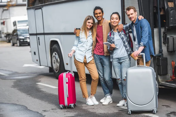 Giovani amici multiculturali felici con borse a ruote in posa vicino autobus di viaggio in strada — Foto stock