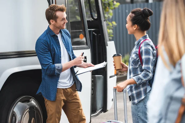 Sorridente masculino ônibus controlador escrito no clipboard enquanto asiático mulher de pé com café xícara perto de ônibus no urbano rua — Fotografia de Stock