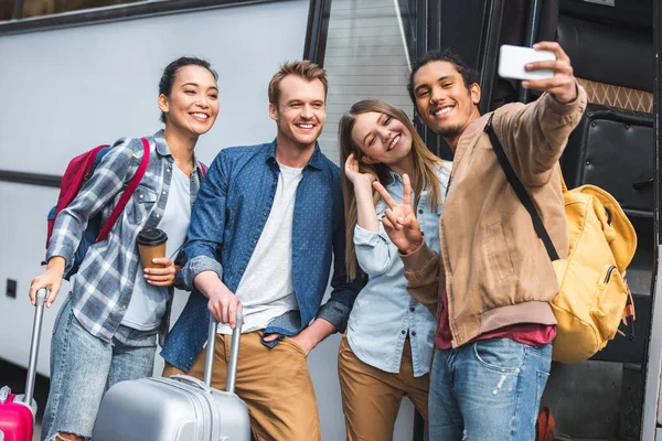 Alegre turista multirracial mostrando señal de paz y tomar selfie con amigos multiculturales en el teléfono inteligente cerca de autobús de viaje en la calle - foto de stock