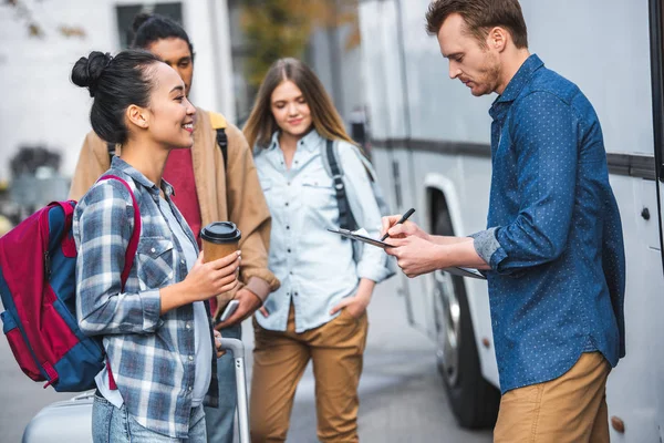 Side view of male travel bus controller writing in clipboard while multicultural tourists standing near bus at street — Stock Photo