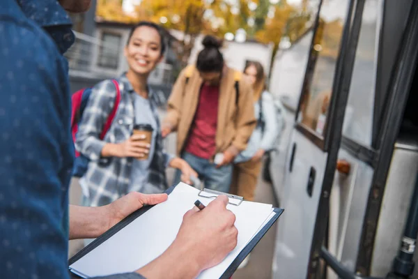 Imagem cortada do controlador de ônibus de viagem escrevendo na área de transferência, enquanto os turistas de pé perto de ônibus na rua da cidade — Fotografia de Stock