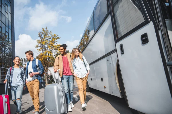 Young smiling interracial couples with wheeled bags walking near travel bus at city street — Stock Photo