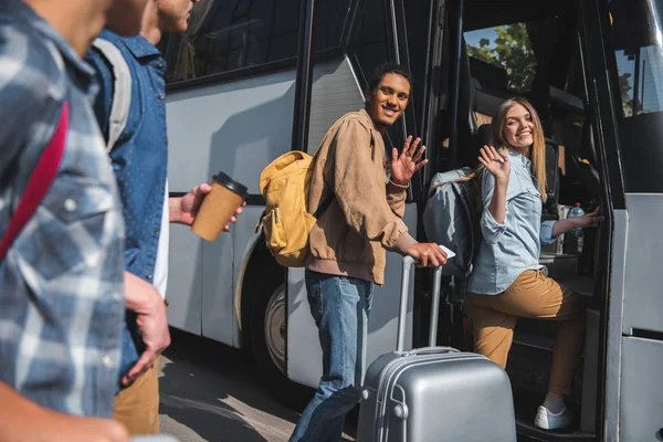 Happy multiethnic tourists with backpacks and wheeled bags waving by hands to friends while walking into bus at street — Stock Photo