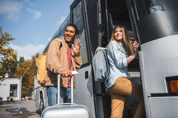 Sonriente turista multirracial masculino con la bolsa de ruedas saludando a mano mientras su novia caminando en autobús de viaje en la calle de la ciudad - foto de stock