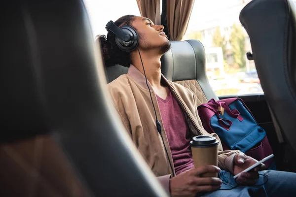 Messa a fuoco selettiva di giovane uomo razza mista in cuffia seduto con tazza di caffè di carta e smartphone in autobus di viaggio — Foto stock