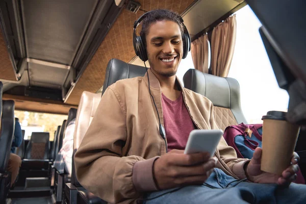 Sorrindo homem de raça mista em fones de ouvido sentado com copo de café de papel e smartphone em ônibus de viagem — Fotografia de Stock