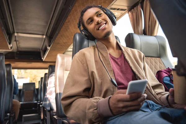 Fröhlicher Mischling mit Kopfhörern, der mit Pappkaffeetasse und Smartphone im Reisebus sitzt — Stockfoto