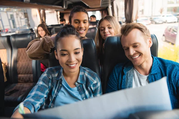 Cheerful multiethnic tourists looking at map during trip on travel bus — Stock Photo