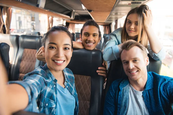 Selective focus of smiling asian woman taking selfie with multicultural friends in travel bus — Stock Photo