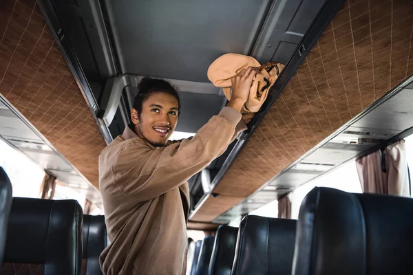 Sonriente joven multirracial hombre poniendo mochila en estante en autobús de viaje - foto de stock