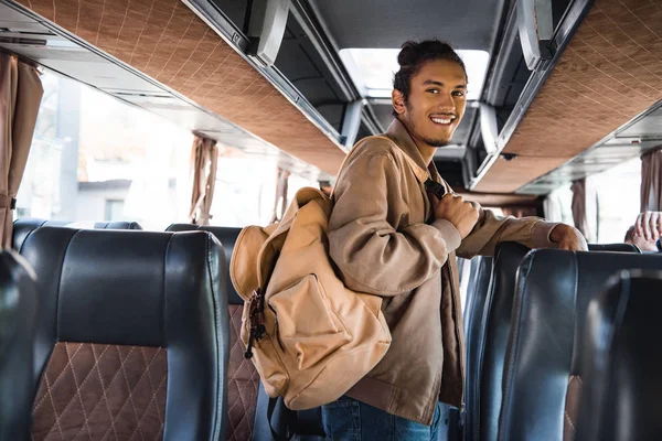 Sonriente turista masculino multirracial con mochila mirando a la cámara en autobús de viaje - foto de stock