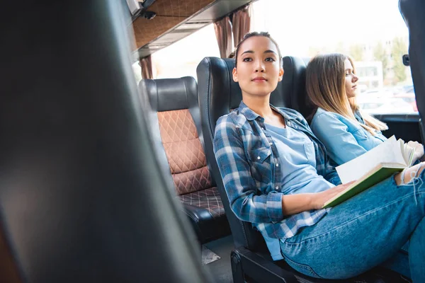 Atractivo asiático mujer leyendo libro mientras su hembra amigos sentado cerca durante viaje en viaje autobús - foto de stock