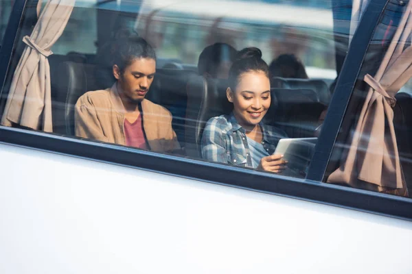 Smiling asian woman using digital tablet while her mixed race boyfriend sitting behind during trip on travel bus — Stock Photo