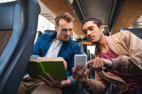 Joven hombre de raza mixta mostrando teléfono inteligente a su amigo varón con libro durante el viaje en autobús de viaje — Stock Photo