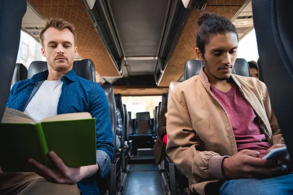 Jeune homme multiracial en utilisant smartphone tandis que son ami masculin livre de lecture pendant le voyage en bus — Photo de stock