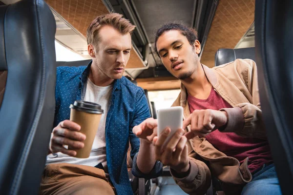 Молодий змішаний чоловік, що показує смартфон своєму чоловікові-друзі з паперовою чашкою кави під час поїздки в автобусі — стокове фото