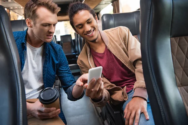 Fröhlicher Mischlingsmensch zeigt seinem männlichen Freund während der Busfahrt sein Smartphone mit Einwegbecher Kaffee — Stockfoto