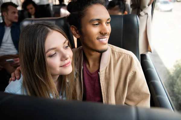 Многонациональная улыбающаяся пара смотрит в окно во время поездки на автобусе — стоковое фото