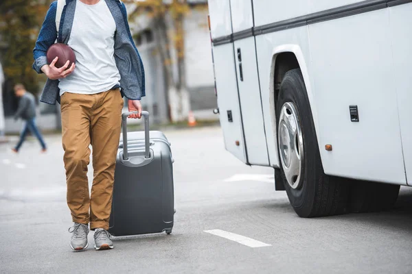 Partial view of man with rugby ball carrying bag on wheels near travel bus at street — Stock Photo