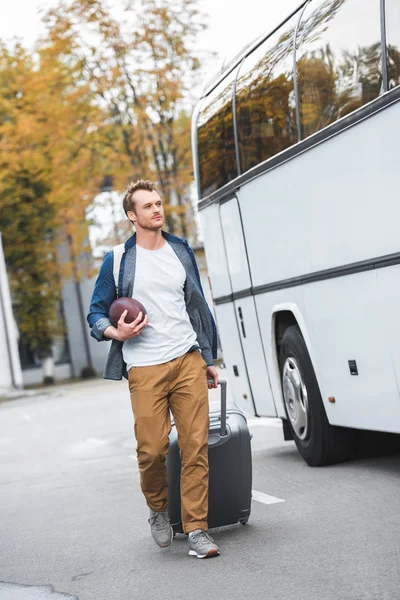Handsome man with backpack and rugby ball carrying bag on wheels near travel bus at street — Stock Photo