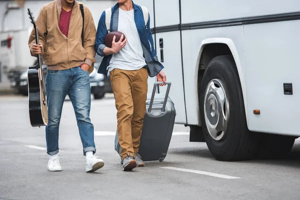 Vista parcial del hombre con la bola de rugby que lleva el bolso de ruedas mientras que su amigo masculino que camina cerca del autobús de viaje en la calle - foto de stock