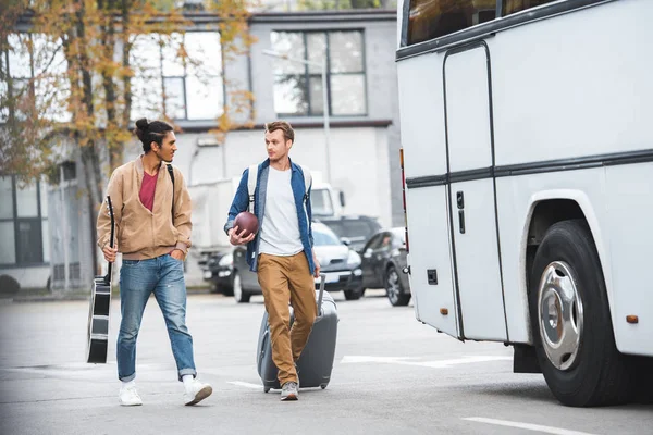 Hombre adulto con pelota de rugby que lleva bolsa de viaje mientras su amigo varón de raza mixta camina cerca de autobús en la calle - foto de stock