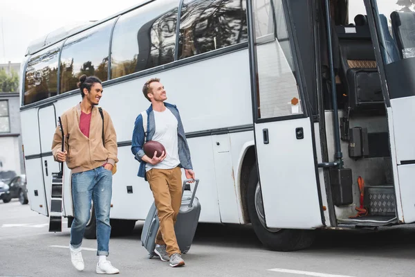 Селективное внимание человека с мячом для регби, несущего сумку с колёсами, в то время как его смешанная раса мужского пола, идущего рядом с автобусом на улице — стоковое фото