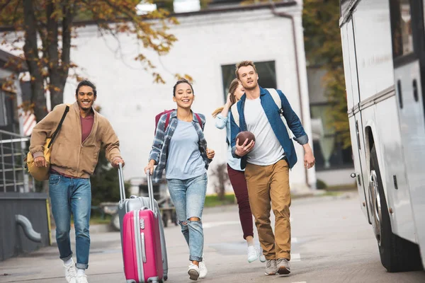 Cheerful young friends with wheeled bags and rugby ball running near travel bus at city street — Stock Photo