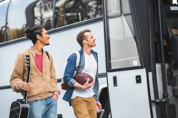 Homme souriant avec balle de rugby portant sac à roulettes tandis que son ami masculin mixte marche près de bus de voyage dans la rue — Photo de stock