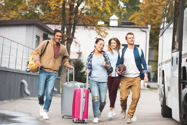 Selective focus of smiling multiethnic tourists with wheeled bags and rugby ball running near travel bus at city street — Stock Photo