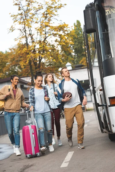 Multiethnic tourists with wheeled bags and rugby ball running near travel bus at city street — Stock Photo