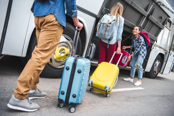 Enfoque selectivo de la mujer sonriente poner la bolsa de ruedas en el autobús de viaje mientras sus amigos caminando cerca de la calle urbana - foto de stock