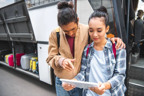 Multirazziale giovane uomo che punta il dito al tablet digitale per asiatica ragazza vicino autobus di viaggio per strada — Foto stock