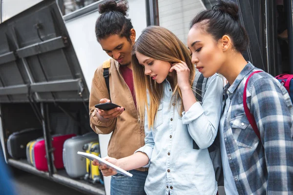 Turistas multiculturales utilizando tableta digital y teléfono inteligente cerca de autobús de viaje en la calle de la ciudad - foto de stock